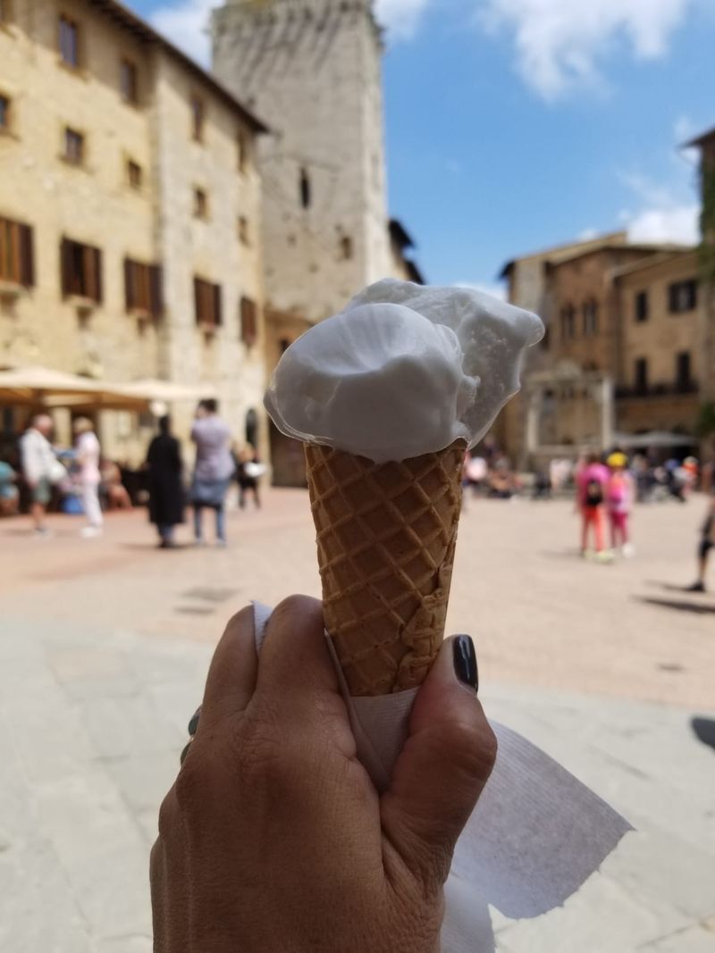 A gelato held up in a Tuscan piazza with medieval towers behind